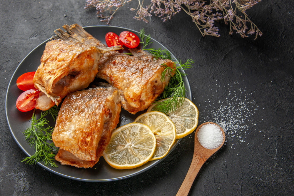 bottom-view-tasty-fish-fry-lemon-slices-cut-cherry-tomatoes-plate-dried-flower-branch-wooden-spoon-black-table